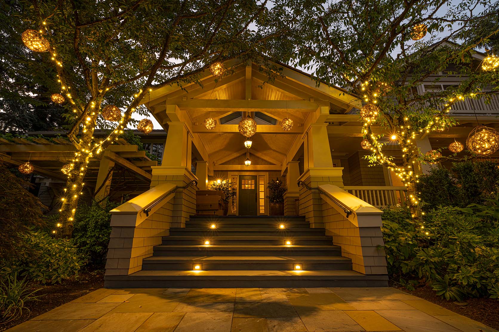 Entry stairs to a house framed by trees wrapped in warm string lights and glowing lanterns at dusk.